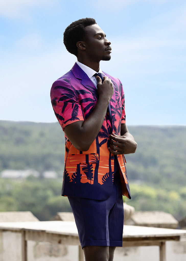 Man standing outdoors wearing summer suit with palm trees on the jacket. Pants is dark Blue and jacket fades from purple at the top to orange at the bottom with a dark blue border.