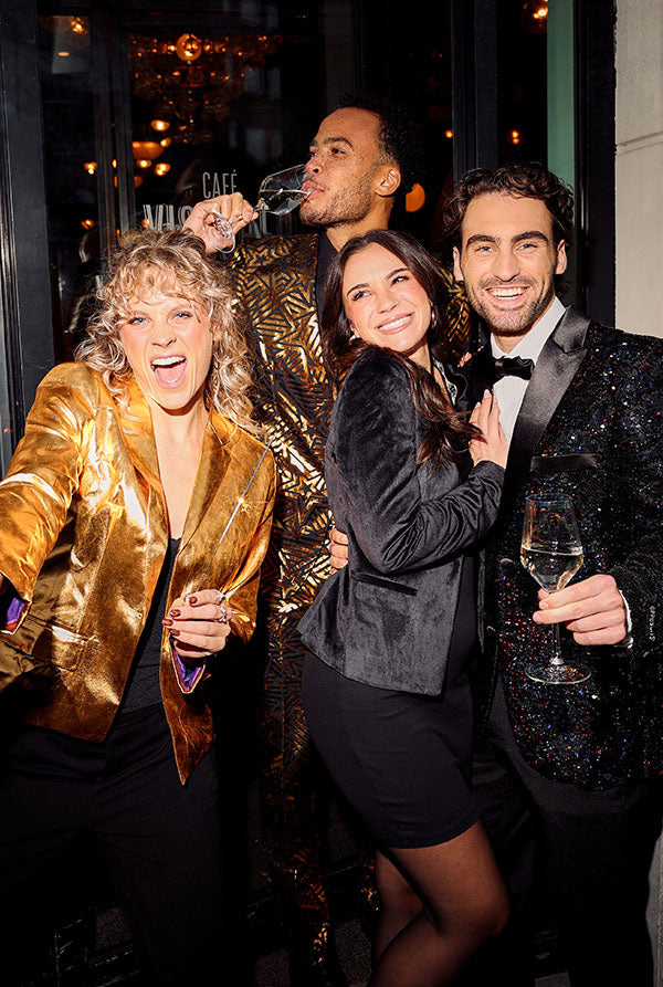 Image with 4 people outside a cafe drinking champagne and wearing OppoSuits NYE outfits. One is wearing a golden blazer, One a black velvet blazer, one a black sequin blazer and one a golden suit with geometric shapes.