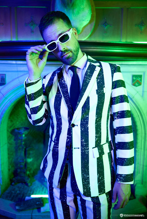 Image of man wearing striped black and white Beetlejuice suit in front of fireplace. Purple green light in the background.