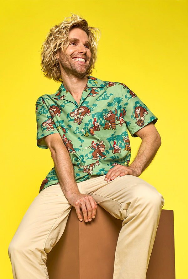 Image with yellow background and man sitting on cardboard box wearing green Donkey Kong Hawaiian and beige pants.  