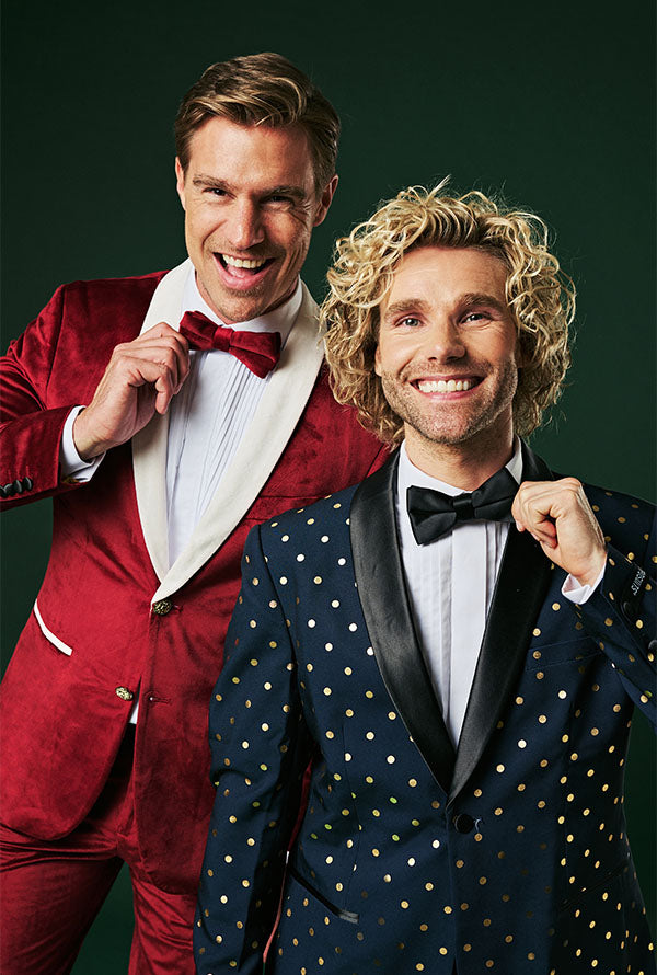 Two men wearing Christmas tuxedos. one is wearing a red one and the other is wearing a blue one.